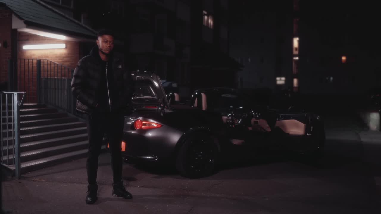 A man in a black puffer jacket stands beside a sports car outside a block of flats at night