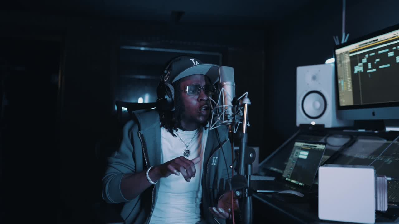 An artist in headphones performs into a studio microphone beside a computer screen showing a music session