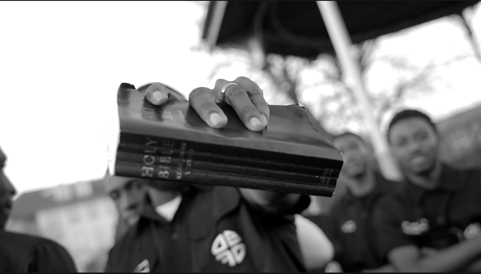 A black-and-white close-up of a hand holding a worn Holy Bible towards the camera, with a group of people behind
