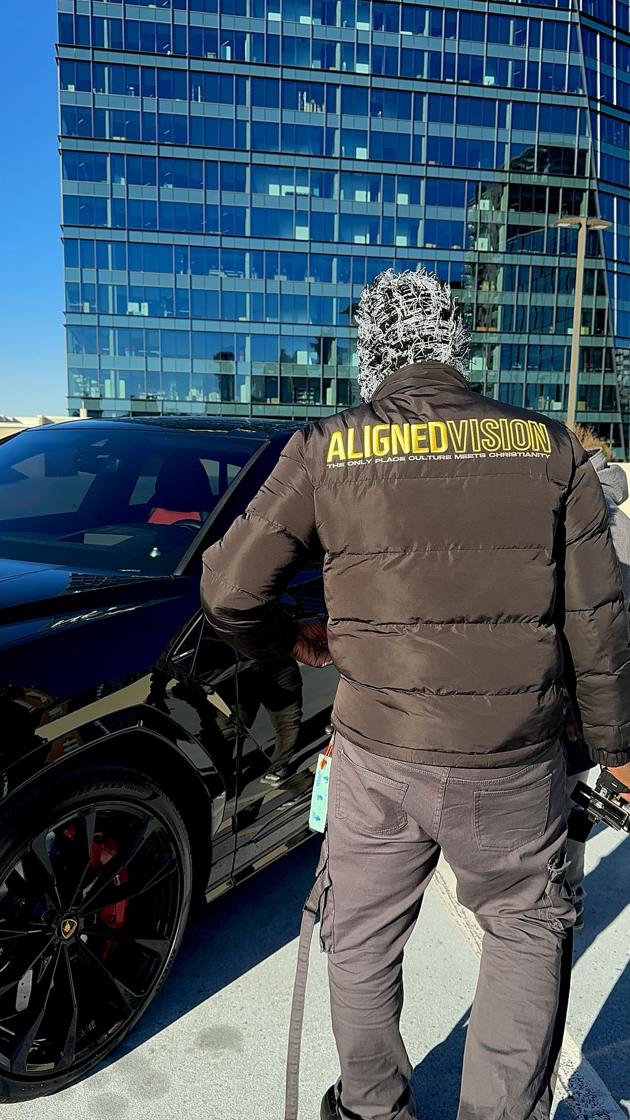 Man seen from behind in a brown Aligned Vision puffer jacket, standing beside a black car in front of a glass office building