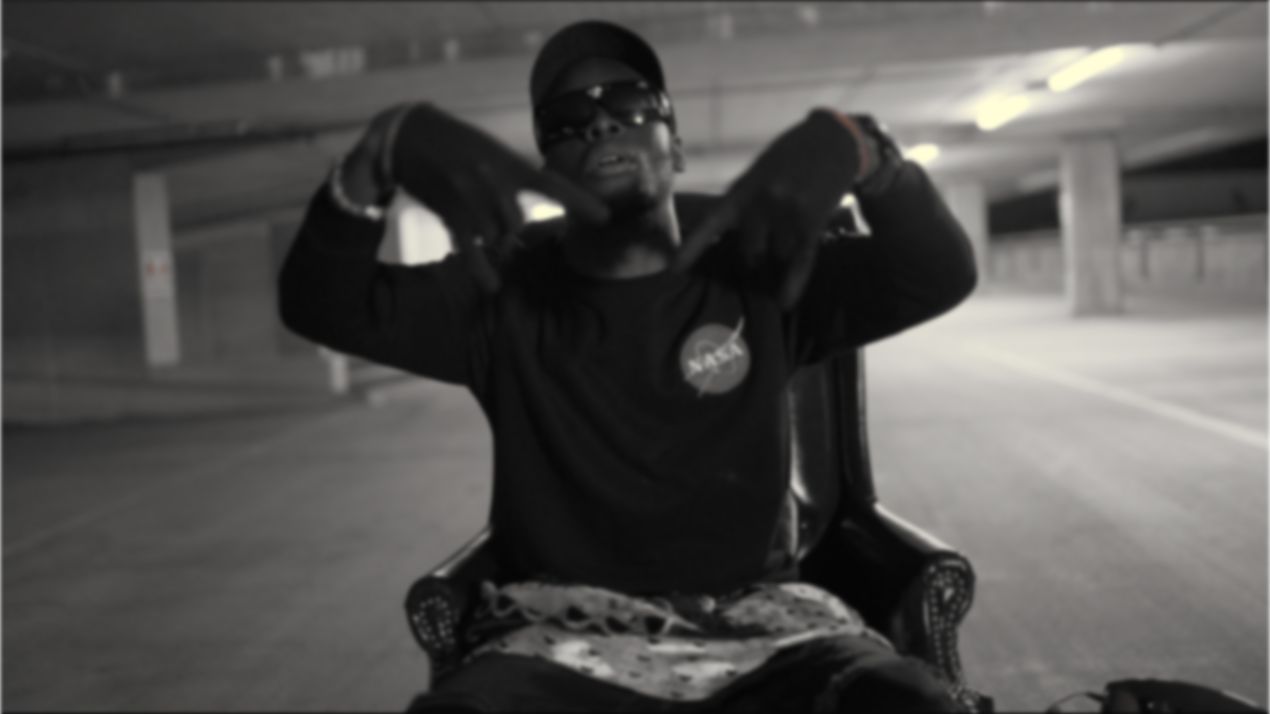 Black and white still of Konola in sunglasses and a cap, seated in an armchair in a car park
