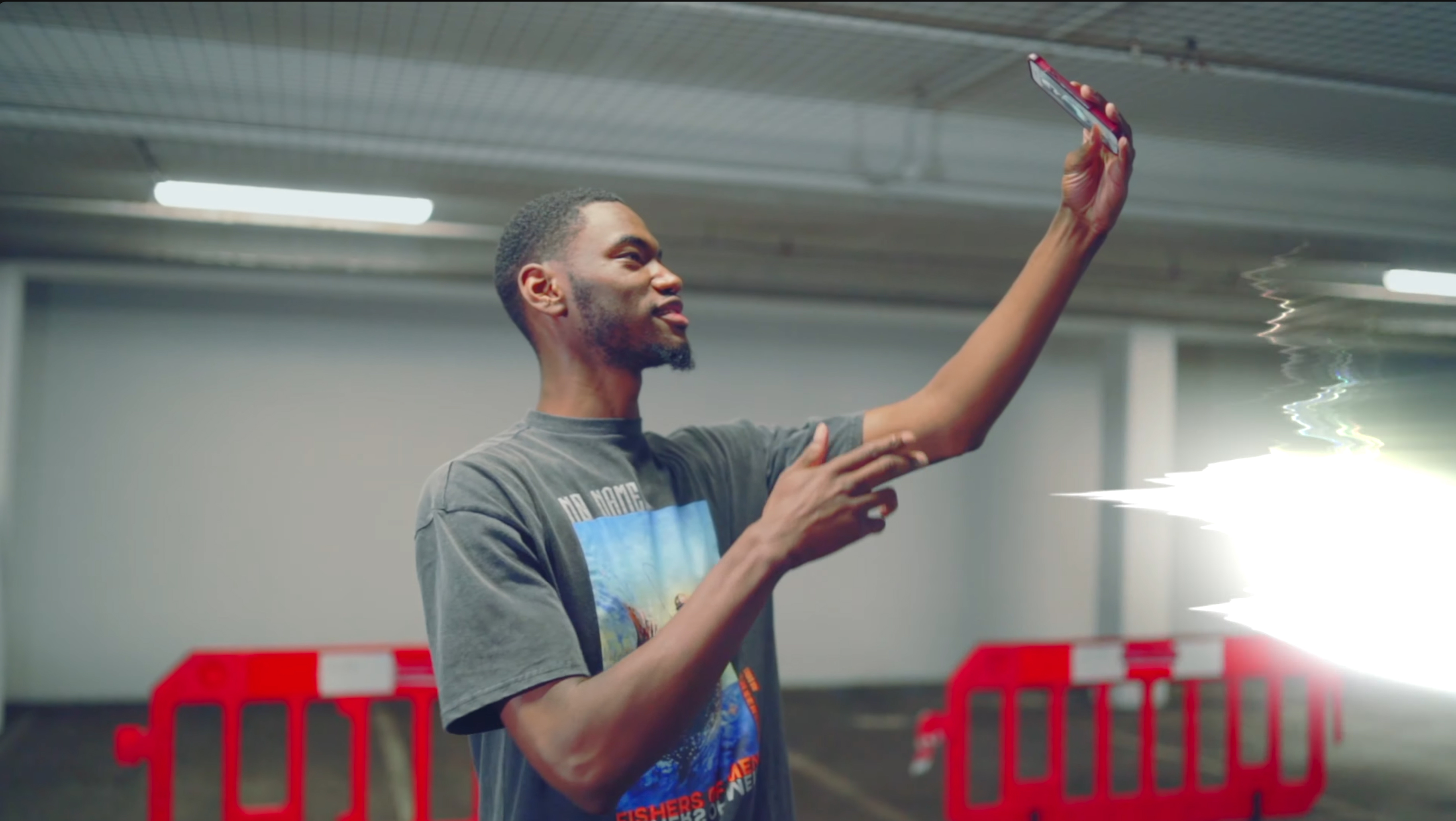 Artist holding up a phone while performing in a car park, with red barriers behind him