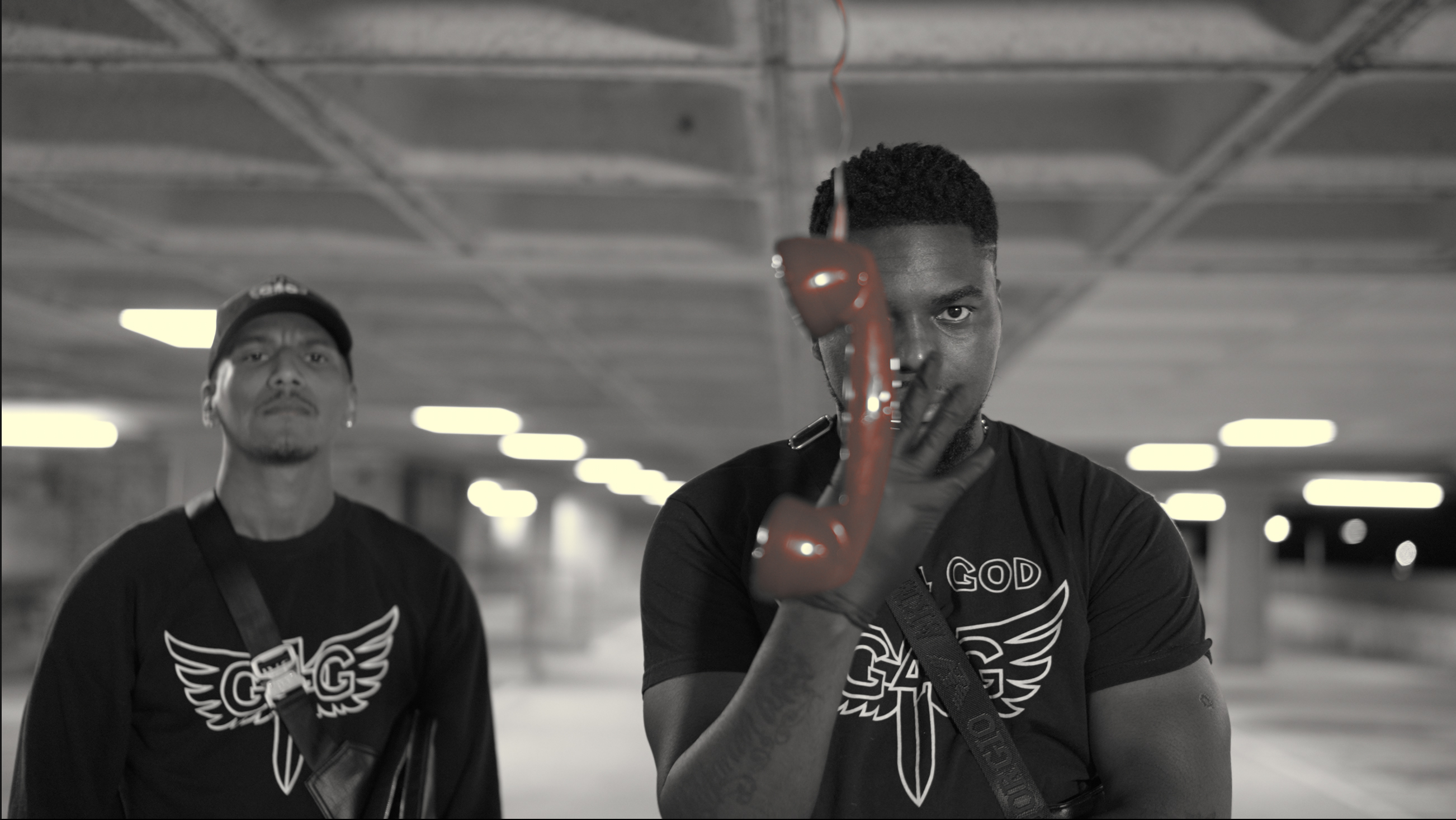 Black and white still of two men in matching winged T-shirts in a car park, one holding a red telephone receiver