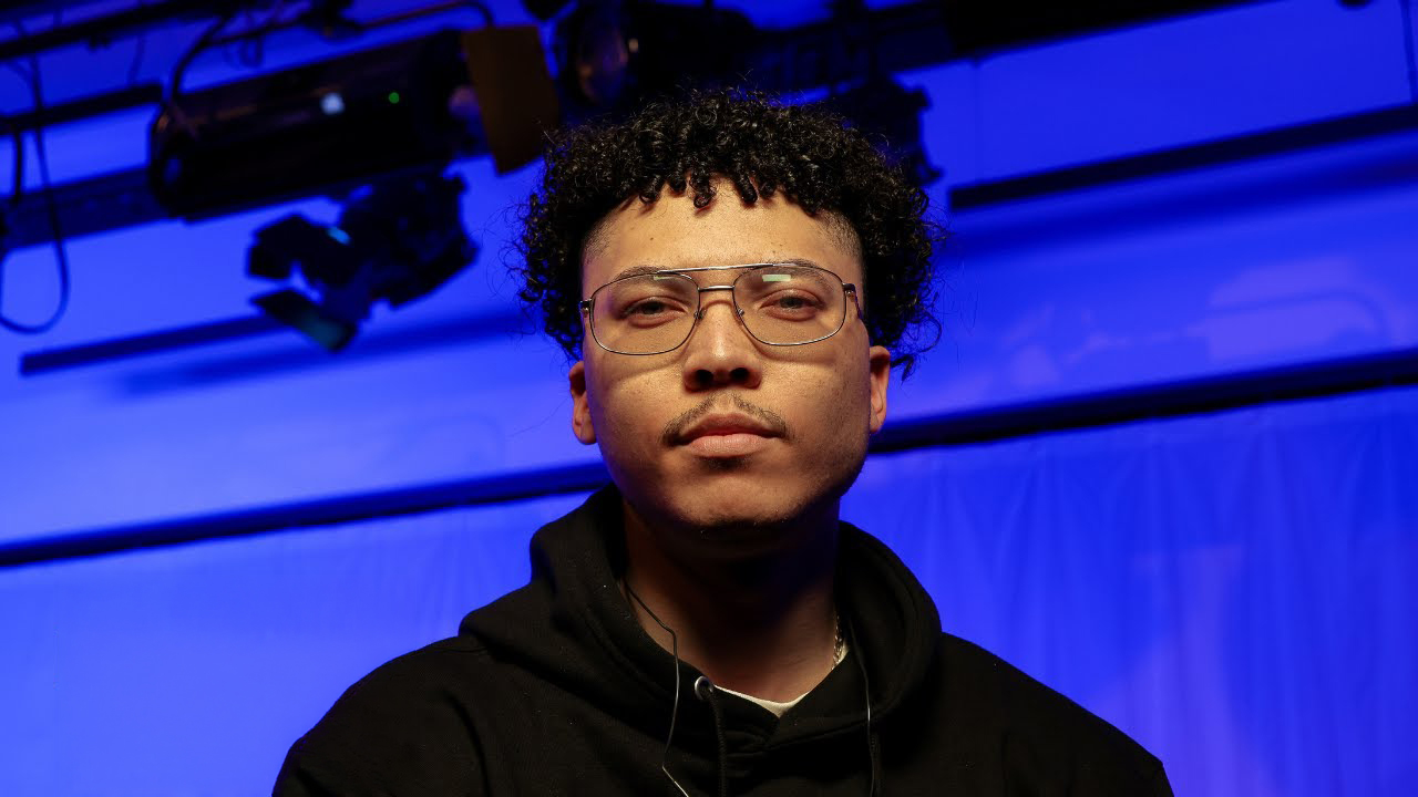 A young man with curly hair, glasses and a black hoodie stands in front of blue stage lighting