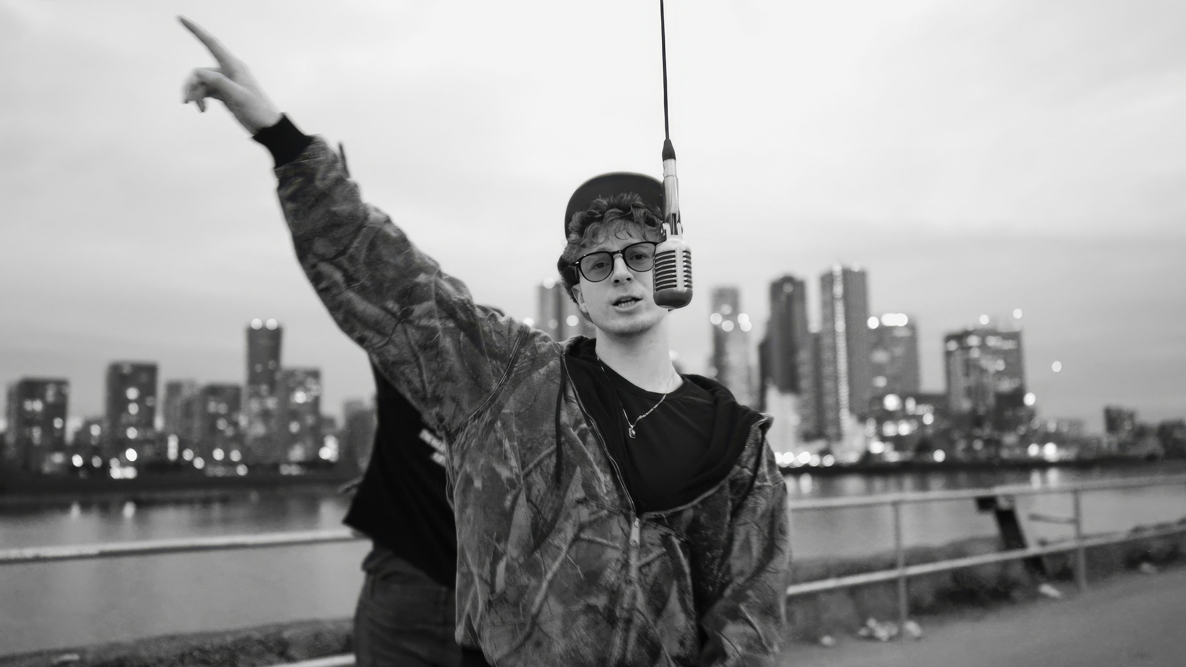 Black and white photo of an artist in glasses and a cap at a hanging microphone by a river, with tower blocks behind