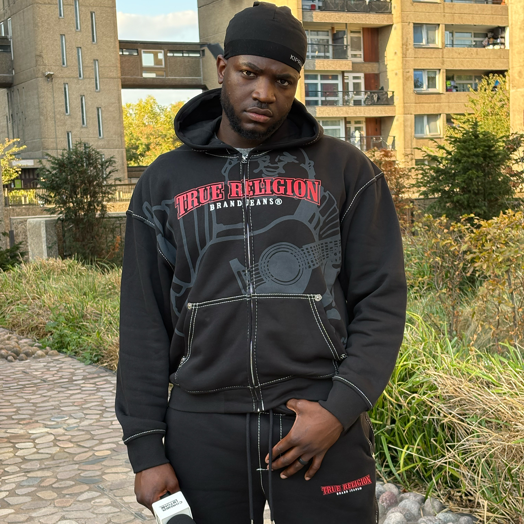 Artist in a black hoodie standing in a garden outside a block of flats