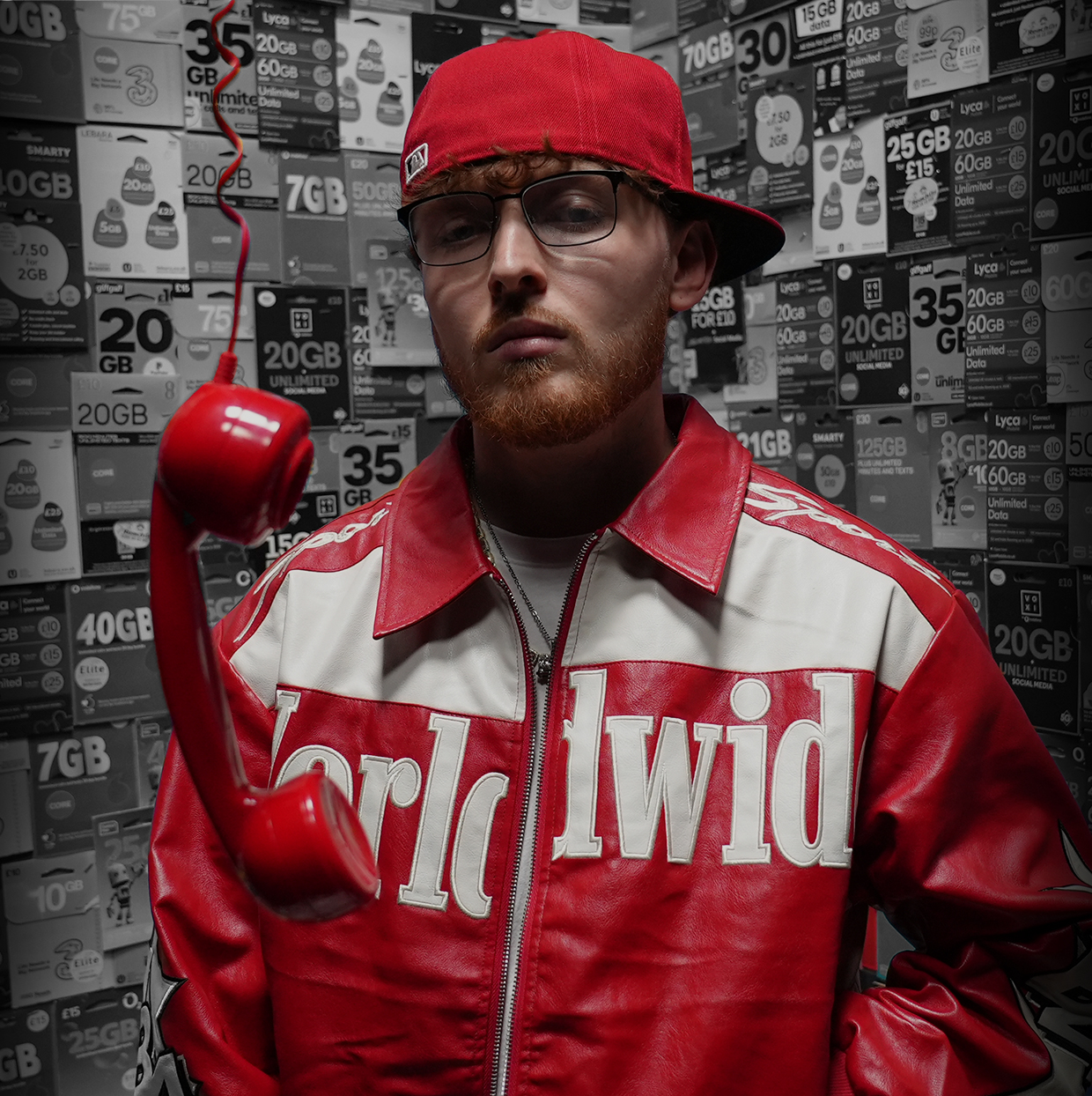 Artist in a red leather jacket and red cap beside a hanging red telephone, in front of a wall of mobile top-up cards