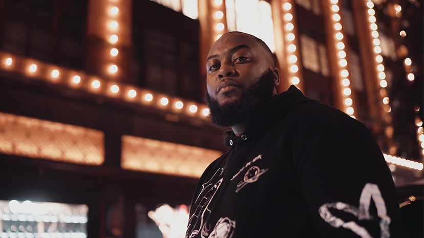 Man in a black hoodie in front of a building lined with warm lights at night