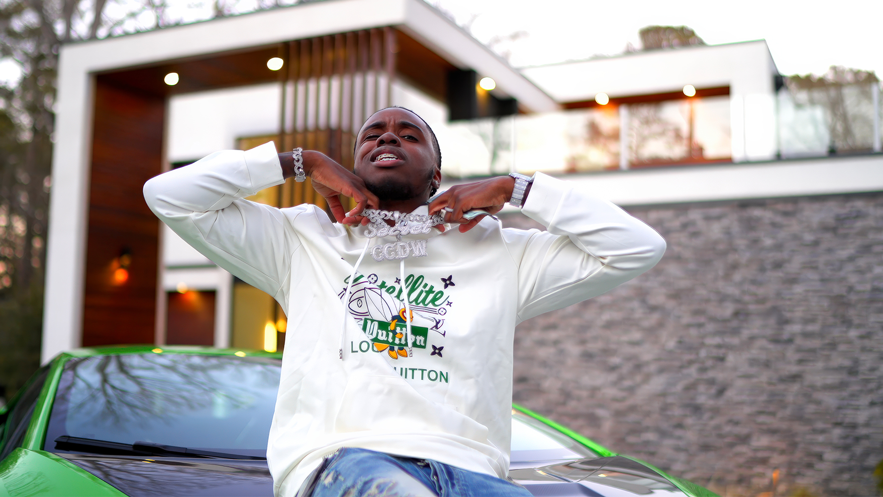 Man in a white hoodie posing on a green car outside a modern house