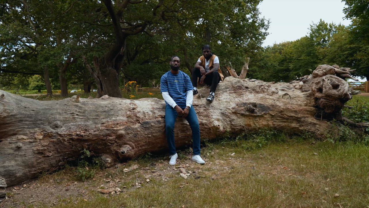 Two men on a large fallen tree trunk in a park, one sitting in front of it and one perched on top