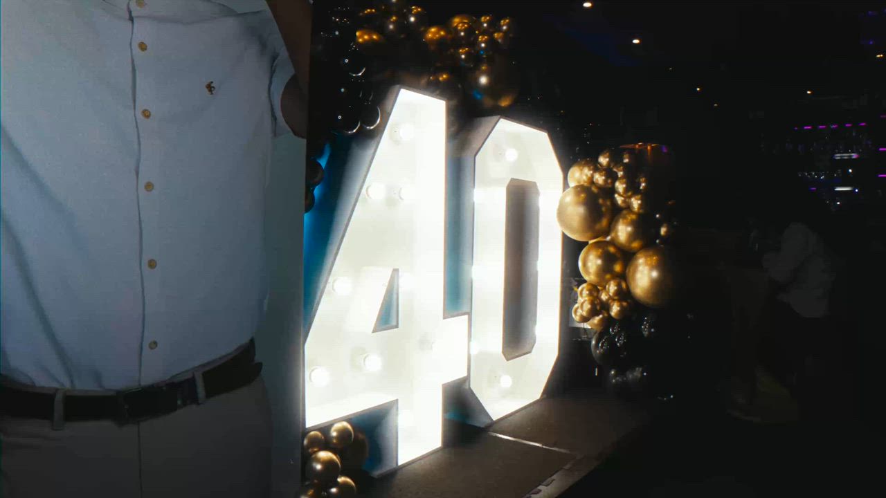 A man in a pale shirt, his face out of frame, standing beside a lit-up number 40 sign and gold balloons at an evening party