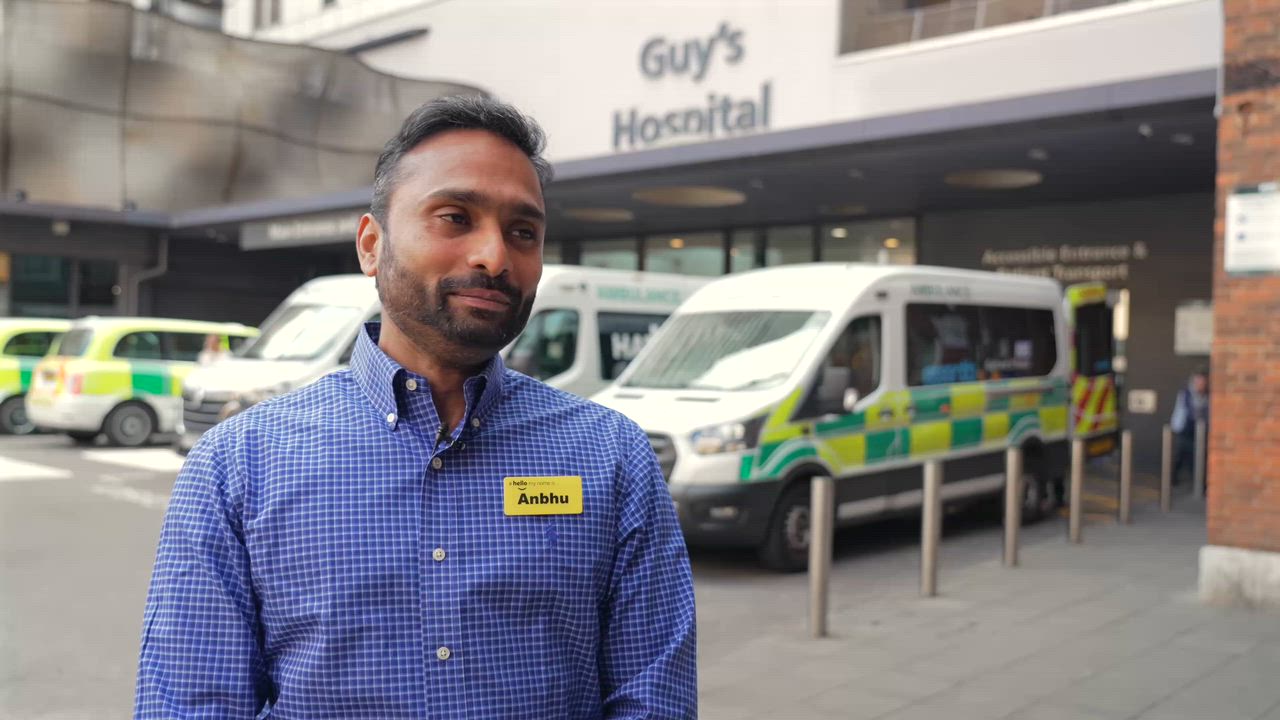 A man in a blue checked shirt and name badge interviewed outside Guy’s Hospital, with ambulances parked behind him